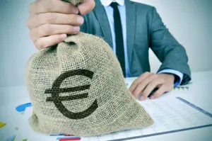 Uomo in giacca solleva un sacco di iuta con simbolo euro, seduto a una scrivania con grafici e tabelle di dati.
