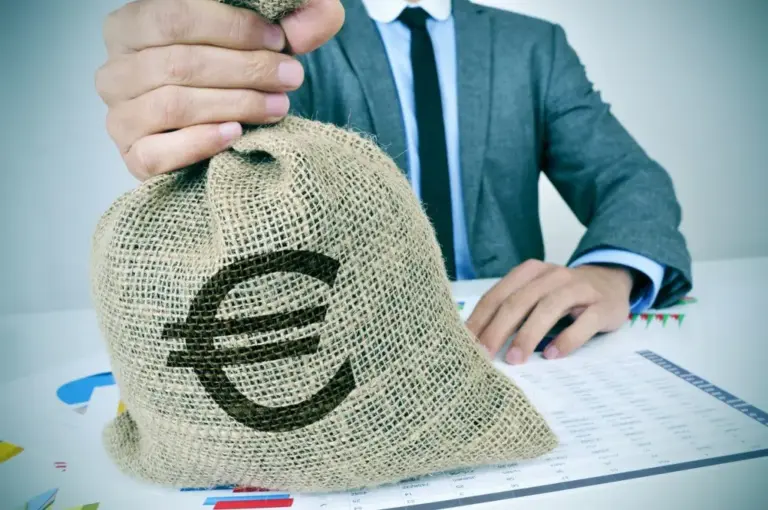 Uomo in giacca solleva un sacco di iuta con simbolo euro, seduto a una scrivania con grafici e tabelle di dati.