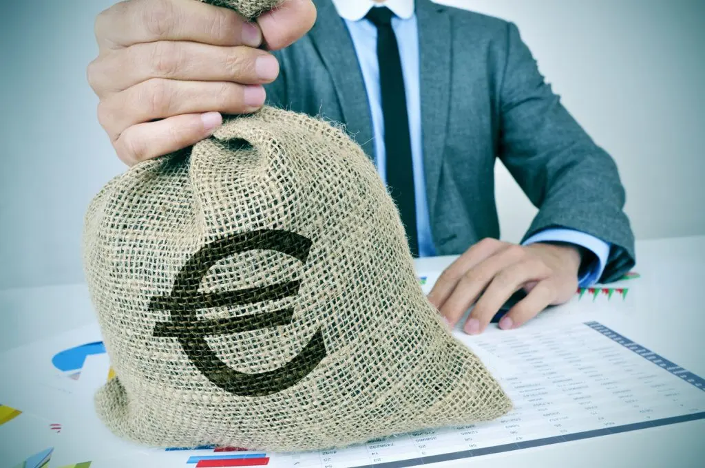 Uomo in giacca solleva un sacco di iuta con simbolo euro, seduto a una scrivania con grafici e tabelle di dati.